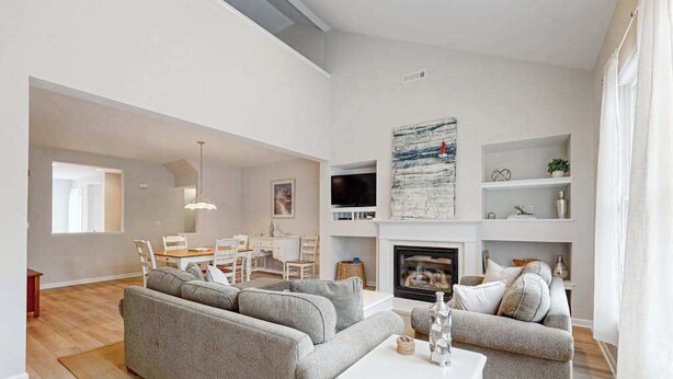 A modern living room with a fireplace, grey sofas, a dining area, and a TV mounted above the fireplace. - Bear Trap Dunes home w/ AC, fireplace, balcony, W/D, beach shuttle (Ocean View)