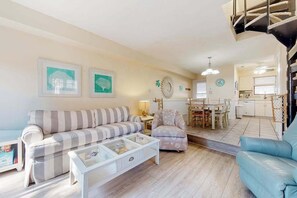 A living room with a sofa, armchair, and coffee table, a dining area with chairs, and a kitchen in the background. - Sea Pines Village - Spectacular condo with W/D, gas grill, balcony, & pool (Bethany Beach)