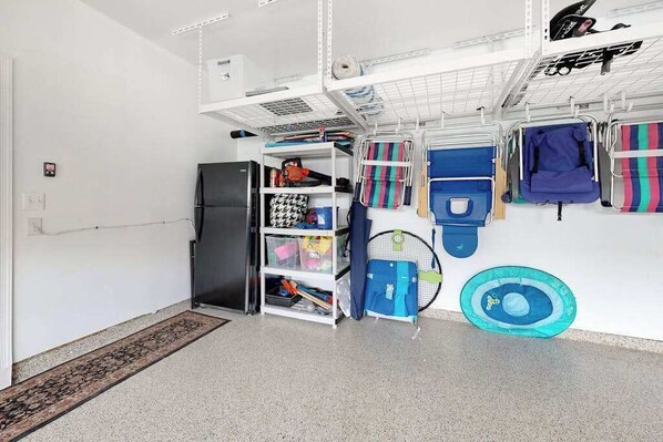 Um quarto com parede branca, geladeira preta, prateleira branca com diversos objetos e tapete azul no chão.