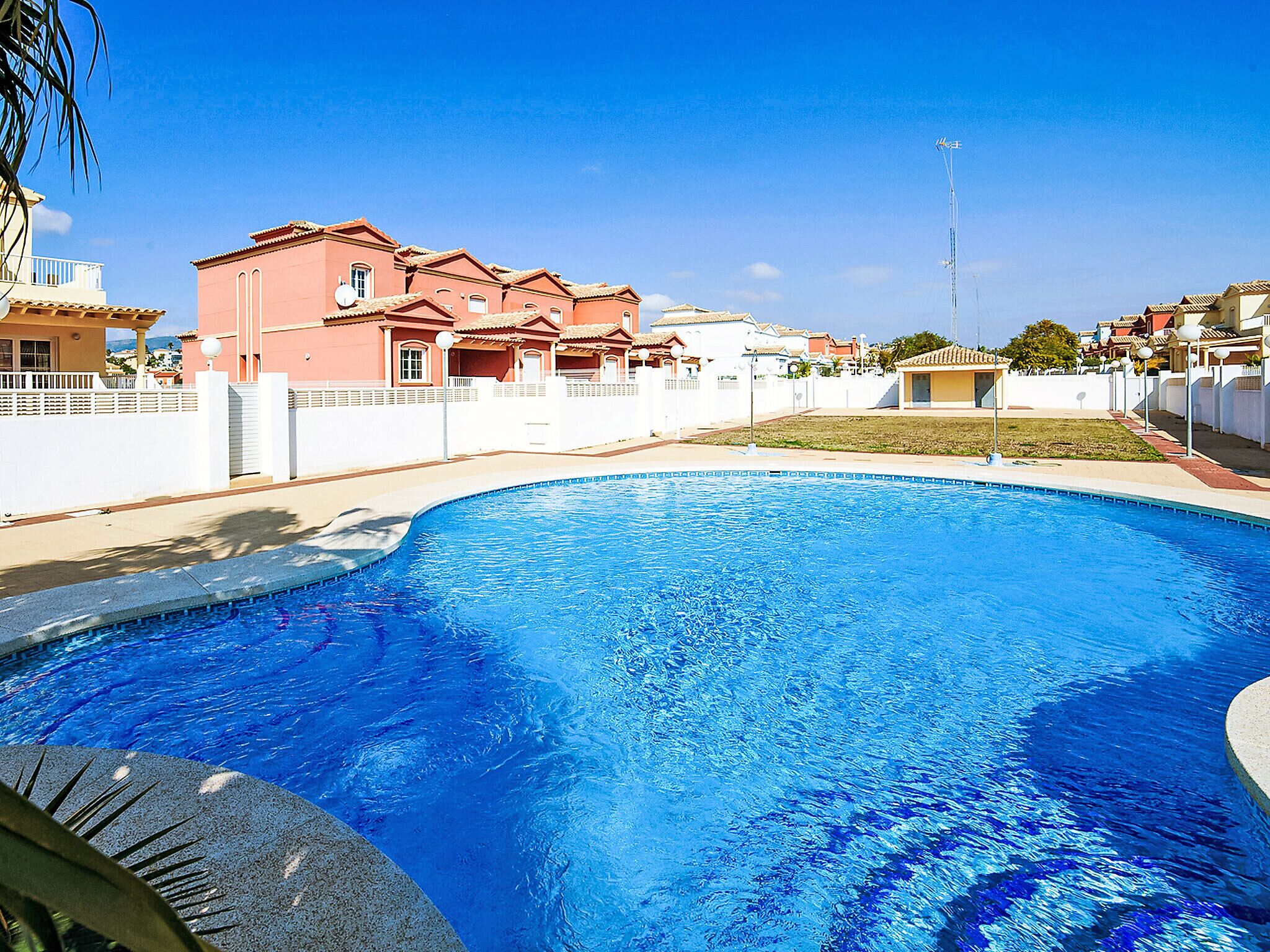 A swimming pool with a clear blue water surface, surrounded by a white fence and a building with a red facade.