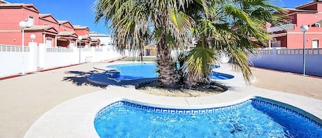 A swimming pool with a palm tree, surrounded by a white fence and buildings with red roofs.
