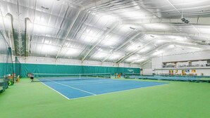 Un court de tennis intérieur doté d’un filet, d’un revêtement de sol vert et d’un plafond haut avec des poutres apparentes.