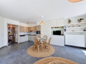 Eine moderne Küche mit weißen Schränken, ein Essbereich mit rundem Tisch und Stühlen sowie ein Fernseher, der über einem Schrank angebracht ist.
