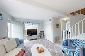 A living room with a sofa, a TV, a chair, and a staircase. - Enchanting condo w/ ocean-view, with balcony, pool, washer/dryer, & dog-friendly (Dewey Beach)
