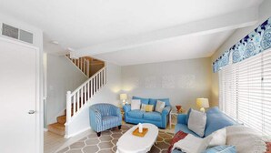 Una sala de estar con una escalera, un sofá azul, un sillón a rayas, una mesa de centro blanca y paredes con diseños.