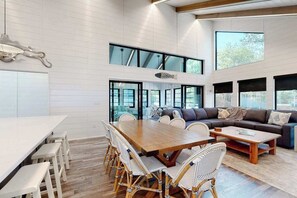 A spacious living area with a wooden dining table, wicker chairs, a sofa, and large windows. - Town of Dewey Beach modern home with grill & outdoor showers - prime location (Dewey Beach)