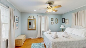 Une chambre à coucher comprenant un lit, un ventilateur de plafond, une table de chevet, une commode et une fenêtre avec des rideaux.