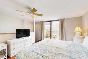Ett hotellrum med en säng, en tv, en takfläkt, ett fönster med gardiner och utsikt över omgivningen.