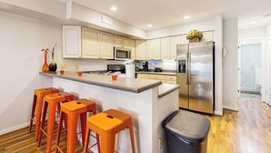 Eine moderne Küche mit weißen Schränken, einem Edelstahlkühlschrank, einer Mikrowelle und einer Bar mit orangefarbenen Hocker.