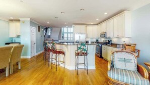 A spacious kitchen with white cabinets, a central island with bar stools, and a dining area with chairs. - Salt Meadows townhouse w/ elevator, gas fireplace & community pool and pier (Fenwick Island)
