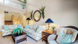 Una sala de estar con un sofá, una mesa de centro, una mesita auxiliar y una silla de mimbre. Se ve una escalera al fondo.