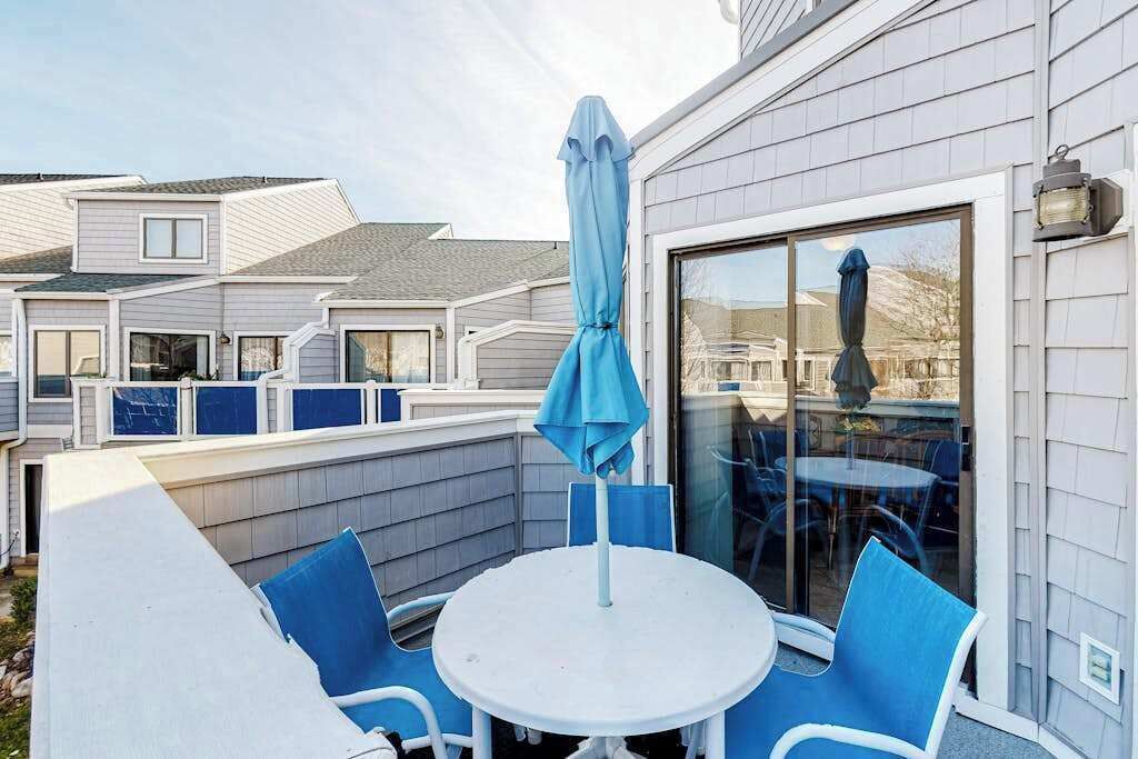 A patio with a round table and blue chairs, a blue umbrella, and a glass door leading to a covered outdoor area.
