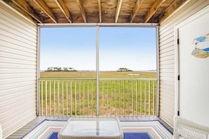 A covered balcony with a view of a grassy field, a wicker table, and a white chair.