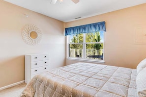 A bedroom with a bed, a dresser, a window with a view of greenery, and a ceiling fan.