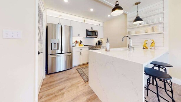 A modern kitchen with white cabinetry, stainless steel appliances, a marble island, and black bar stools. - Sea Colony ground floor condo, updated with fast WiFi, gas fireplace, & deck (Bethany Beach)