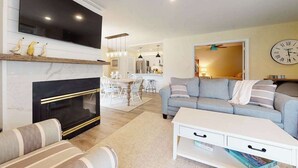 A living room with a fireplace, a television, a sofa, and a dining area in the background. - Sea Colony ground floor condo, updated with fast WiFi, gas fireplace, & deck (Bethany Beach)