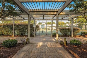 A covered outdoor area with a wooden pergola, paved walkway, and surrounding greenery.