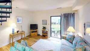 A living room with a sofa, a TV, a wooden coffee table, a bookshelf, and a staircase. - Sea Pines Townhouse Steps From the Water w/Free WiFi, Shared Pool, Private W/D, AC (Bethany Beach)