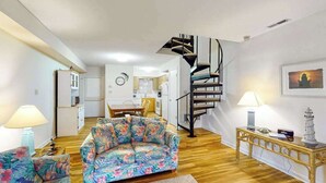 A living room with a sofa, a table, a lamp, a staircase, and a kitchen in the background. - Sea Pines Townhouse Steps From the Water w/Free WiFi, Shared Pool, Private W/D, AC (Bethany Beach)
