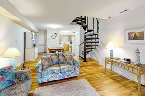 A living room with a sofa, a table, a lamp, a staircase, and a kitchen in the background. - Sea Pines Townhouse Steps From the Water w/Free WiFi, Shared Pool, Private W/D, AC (Bethany Beach)