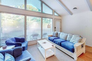 A spacious living room with large windows, a white sofa, a blue sofa, and a white coffee table.