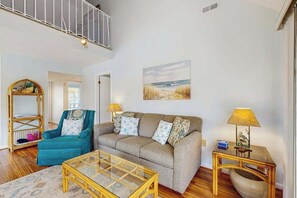 A living room with a sofa, a glass coffee table, a chair, a lamp, and a painting on the wall. - Top-floor Sea Colony gem with AC & fast WiFi - walk to beach, pools & tennis (Bethany Beach)