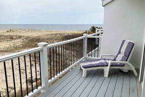 A balcony with a white railing, a single lounge chair with a cushion, and a view of a beach and ocean.