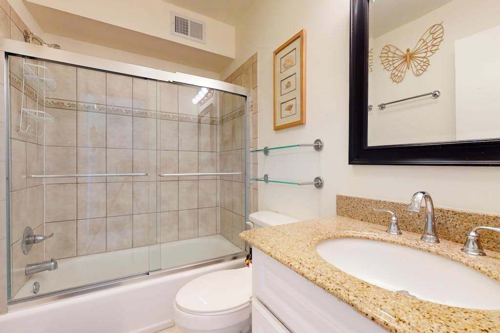 A bathroom with a glass-enclosed shower, a white toilet, a granite countertop sink, and a wall-mounted mirror.