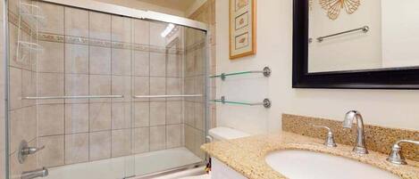 A bathroom with a glass-enclosed shower, a white toilet, a granite countertop sink, and a wall-mounted mirror.