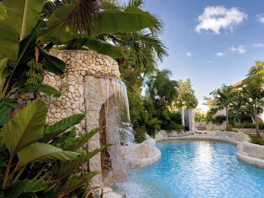 A pool with a waterfall, surrounded by tropical plants and a stone wall.