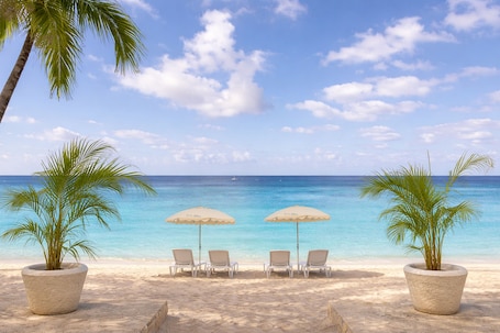 A beach with two palm trees, two beach chairs, and two beach umbrellas.