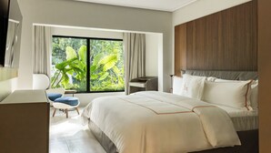 A modern hotel room with a large bed, a desk, a chair, and a view of lush greenery through the window.