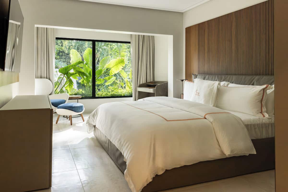Ein modernes Hotelzimmer mit einem großen Bett, einem Schreibtisch, einem Stuhl und Blick auf üppiges Grün durch das Fenster.