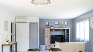 A modern living room with a sofa, a dining table with chairs, a television, and a wall-mounted shelf with decorative items. - Gîte Le Grand Chêne: Shared Pool, Wi-Fi & Air Conditioning in Lesperon (Lesperon)