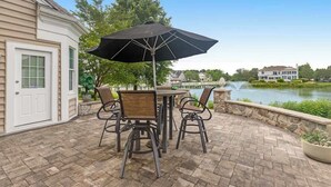 Patio esterno con tavolo e sedie, un ombrellone nero e vista su un lago e case sullo sfondo.