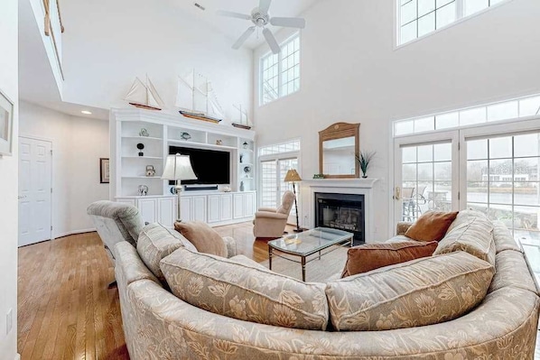 A spacious living room with a large sofa, a flat-screen TV, a fireplace, and a ceiling fan. - Bethany Lakes home with waterfront views, pool access, tennis, & private patio (Bethany Beach)