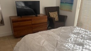 A bedroom with a bed, a TV on a wooden dresser, a chair, and a painting on the wall.