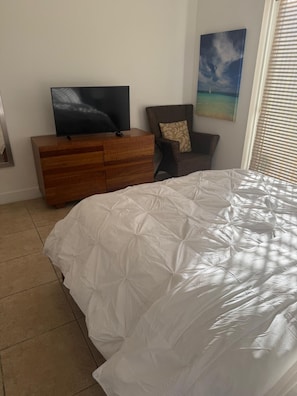 Ein Schlafzimmer mit einem Bett, einem Fernseher auf einer Holzkommode, einem Sessel und einem Bild an der Wand.