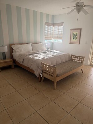 Un dormitorio con cama, mesita de noche, silla, ventilador de techo y ventana con persianas.