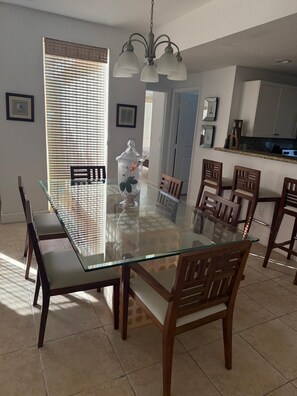 Un comedor con una mesa de vidrio, sillas de madera, una araña y una cocina al fondo.