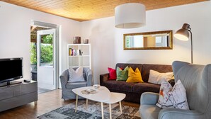 A modern living room with a flat-screen TV, grey sofas, a white coffee table, and a bookshelf.