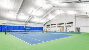 Een binnentennisbaan met een net, blauw-groene vloer en een hoog plafond met zichtbare balken.