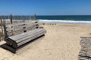 Un banco de madera en una playa arenosa con una cerca de madera al fondo y el océano en la distancia.