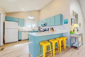 Una cocina con gabinetes color turquesa, un refrigerador blanco, un microondas y una isla central con taburetes amarillos.