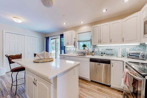 A modern kitchen with white cabinets, a central island, a dishwasher, an oven, and a window with curtains. - The Reservation - dog-friendly home with fenced yard, deck, grill, & beach gear (Ocean View)