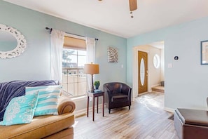 A room with a sofa, a chair, a lamp, a window with curtains, and a door leading to another room. - The Reservation - dog-friendly home with fenced yard, deck, grill, & beach gear (Ocean View)