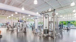 Une salle de sport spacieuse, dotée de divers appareils d’exercice, d’un plafond haut avec un éclairage industriel et de grandes fenêtres laissant entrer la lumière naturelle.