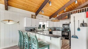 Una cocina con una isla central, taburetes de bar, armarios blancos, techo de madera y vistas al exterior.
