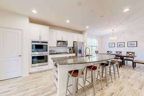 Una cocina moderna con armarios blancos, electrodomésticos de acero inoxidable, una isla de granito con taburetes y un comedor con sillas de madera.