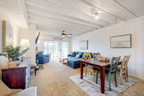 A spacious living area with a wooden ceiling, a dining table with chairs, a blue sofa, and a small table with a lamp and plant.